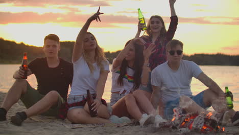 The-young-colleagues-are-sitting-in-around-bonfire-on-the-lake-coast.-They-are-dancing-with-beer-and-talking-to-each-other-at-sunset.